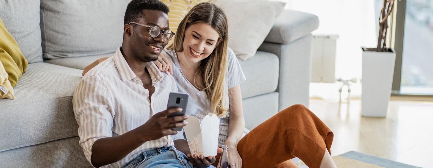 a man and woman with take out, sit against a couch on their living room floor looking at their cell phone
